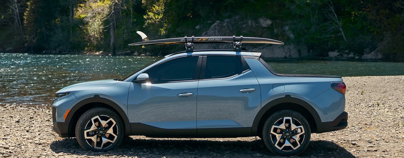 Side view of a blue 2024 Hyundai Santa Cruz parked at the beach with a surfboard on the roof.